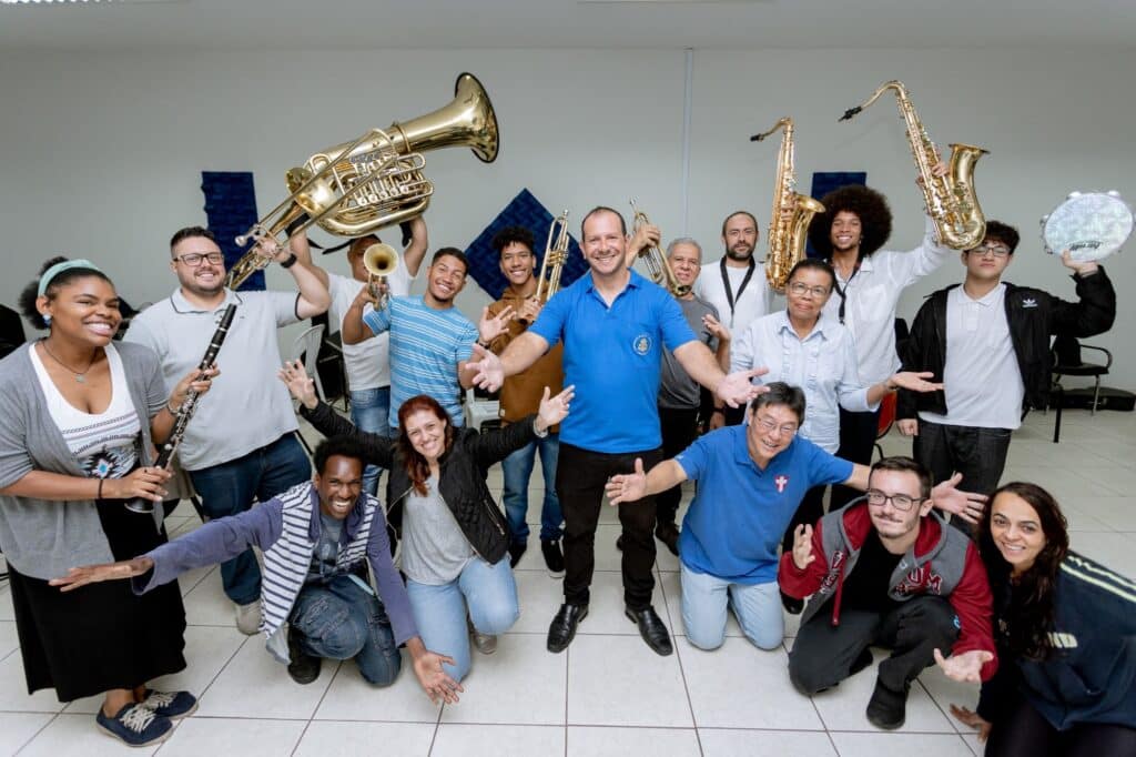 A imagem mostra um grupo grande e animado de músicos, muitos segurando seus instrumentos, sorrindo para a câmera. Há uma mistura de instrumentos de sopro, incluindo trompetes, saxofones, e um tuba. Um homem de camisa azul está no centro da foto, com os braços abertos, como se estivesse conduzindo ou liderando o grupo. As pessoas ao redor dele parecem ser de diversas idades e etnias. A atmosfera é descontraída e festiva, sugerindo uma reunião de banda ou um ensaio. O fundo é simples, uma parede branca com um pouco de decoração, indicando um ambiente interno como um salão de ensaio ou uma sala de aula. A iluminação é uniforme, sem sombras fortes, contribuindo para a clareza da imagem.