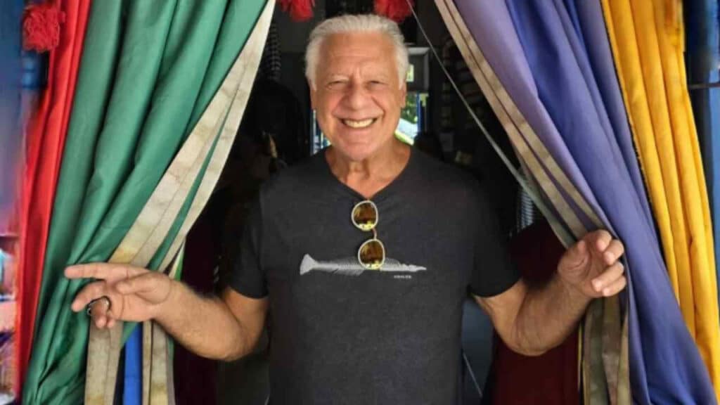 Antonio Fagundes com óculos preso na camiseta, camiseta preta e sorrindo com braços abertos