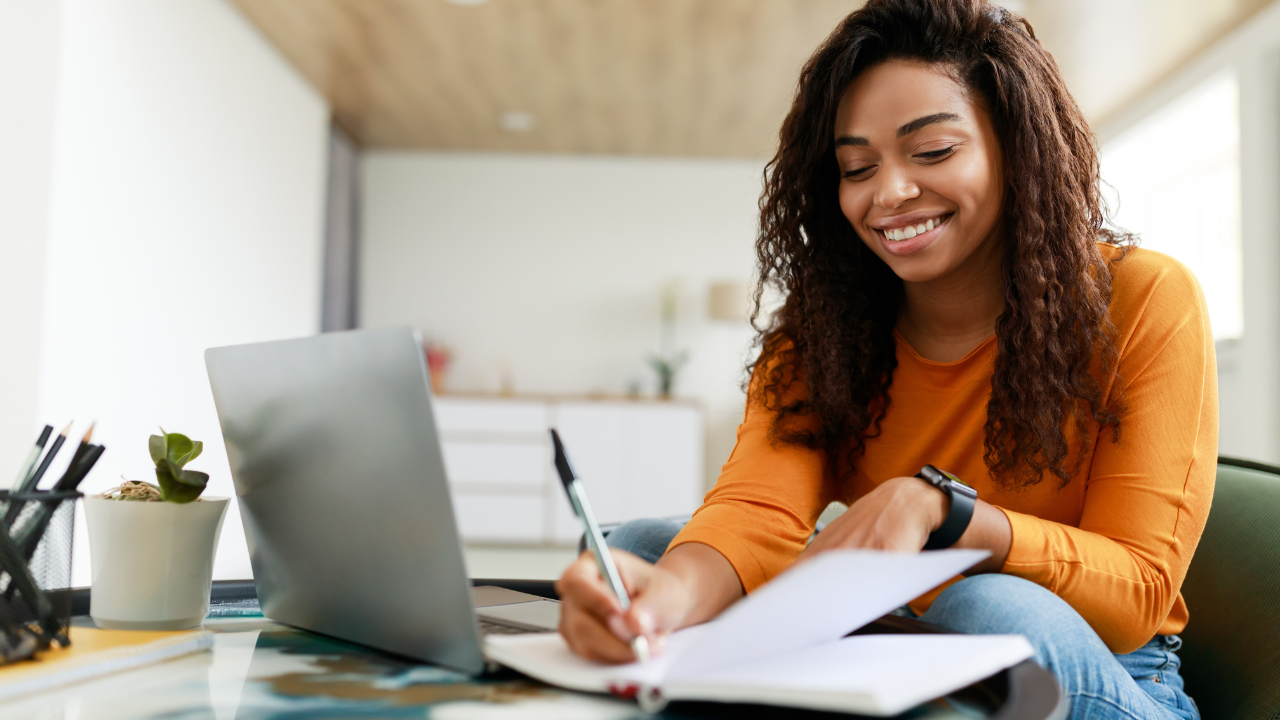 Uma mulher negra de cabelos longos e castanhos sorri enquanto escreve em um caderno aberto com uma caneta. Ela está sentada em um ambiente interno, que parece ser um escritório ou sala de estar bem iluminada, vestindo uma blusa laranja e calça jeans. Um laptop prateado está aberto na mesa à sua frente, ao lado de um pote com lápis e uma pequena planta.