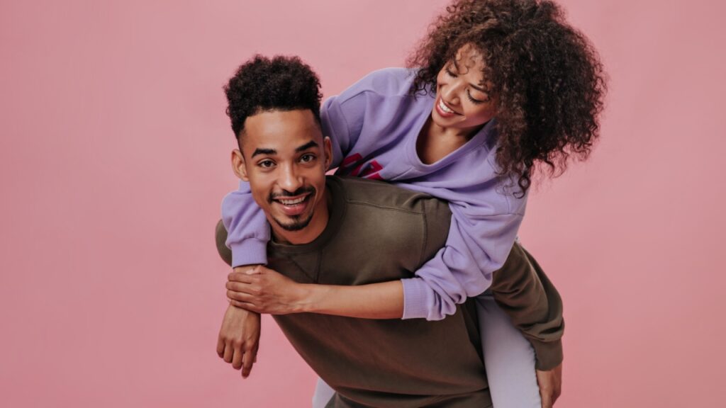 uma mulher negra está nas costas de um homem negro e os dois sorriem em fundo rosa