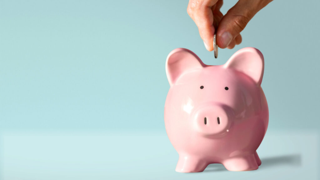 mão colocando uma moeda em um cofre de porquinho rosa em fundo azul