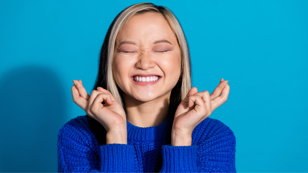 Mulher jovem sorri amplamente com os olhos fechados, demonstrando alegria e expectativa. Ela veste um suéter azul e mantém as mãos erguidas próximas ao rosto, com os dedos cruzados em gesto de torcida. O fundo é azul liso, destacando a expressão positiva e confiante.