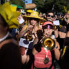 A imagem mostra um grupo de pessoas participando de um desfile de Carnaval. Algumas pessoas estão tocando trompetes, enquanto outras usam fantasias coloridas e acessórios divertidos. Uma mulher usa um chapéu grande e amarelo, enquanto outra tem óculos escuros e uma camiseta sem mangas. O ambiente é festivo, com muitos participantes sorrindo e se divertindo.