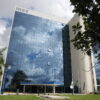 Imagem de um edifício de vários andares com fachada de vidro refletivo, onde se pode ver o céu e nuvens refletidos. O prédio é identificado pelo logo do INSS na parte superior. Na frente, há uma área ajardinada com algumas árvores e bandeiras.