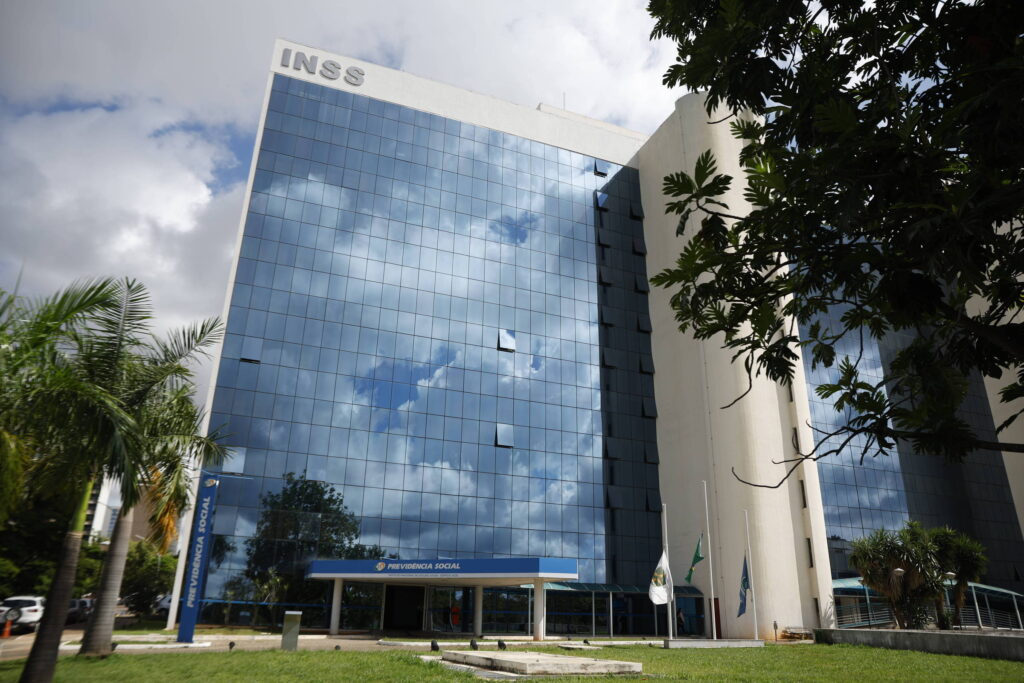 Imagem de um edifício de vários andares com fachada de vidro refletivo, onde se pode ver o céu e nuvens refletidos. O prédio é identificado pelo logo do INSS na parte superior. Na frente, há uma área ajardinada com algumas árvores e bandeiras.