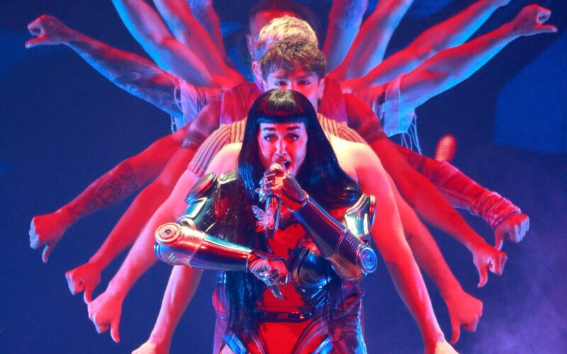 Uma artista se apresenta no palco, vestindo um traje vermelho e azul, com detalhes metálicos. Atrás dela, várias pessoas estão posicionadas, com os braços estendidos, criando uma ilusão de múltiplos braços. A iluminação é colorida, com tons de azul e vermelho.