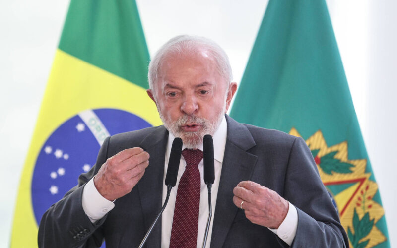 Homem de terno e gravata fala em microfones com bandeiras do Brasil e outra institucional ao fundo.