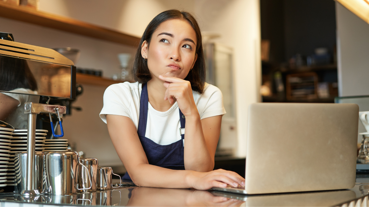 Mulher asiática de cabelo curto e avental com cara pensativa em um balcão de cafeteria.