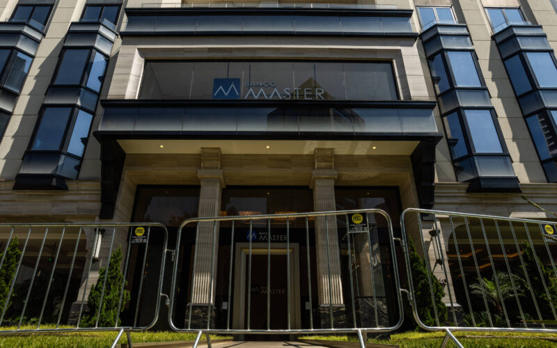 Entrada de prédio com colunas e vidro, protegida por barreiras metálicas alinhadas na calçada. Fachada moderna com janelas espelhadas e o nome