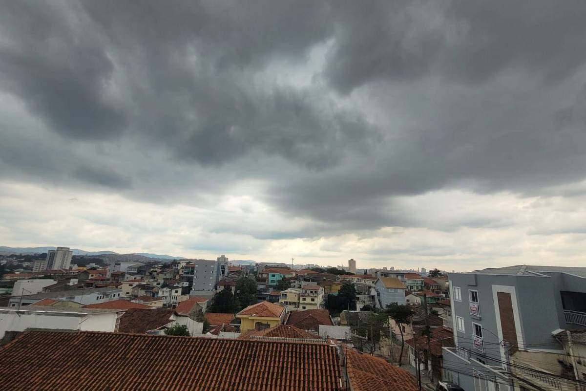 Vista panorâmica de bairro urbano com casas de telhado vermelho e prédios baixos sob céu carregado de nuvens escuras, indicando tempo nublado.