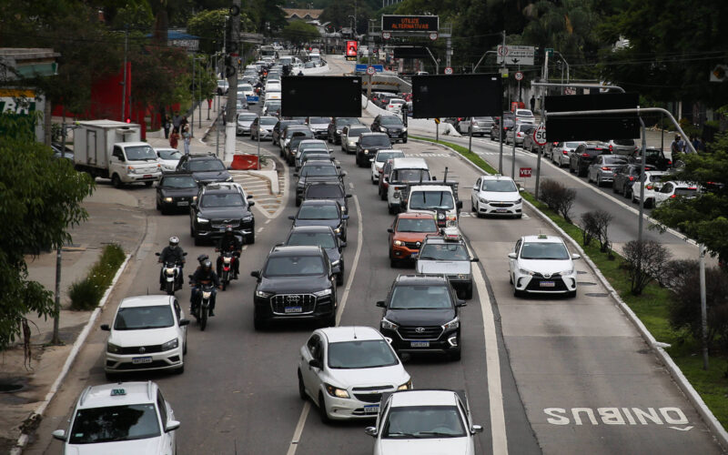 Vários veículos, incluindo carros e motos, formam congestionamento em avenida de pista dupla com faixa exclusiva para ônibus à direita. Árvores e construções cercam a via em dia nublado.