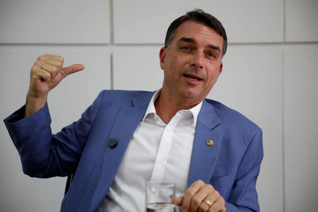 Homem de meia-idade veste terno azul e camisa branca, sentado em ambiente interno com parede branca ao fundo. Ele gesticula com a mão esquerda apontando para trás e segura um copo com a mão direita.