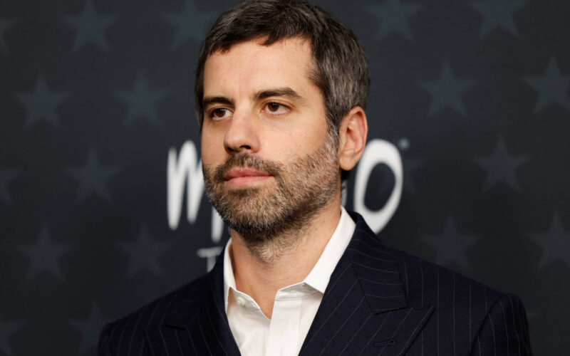 Homem de cabelos curtos e barba aparada veste terno escuro listrado e camisa branca, posando diante de fundo escuro com estrelas e texto desfocado.