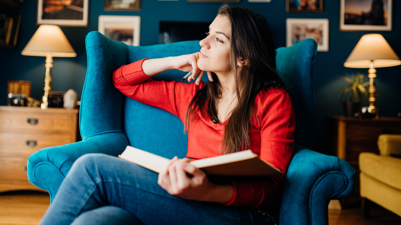 Uma mulher usando camiseta vermelha, segurando um livro, sentada no sofá e olhando para a janela.