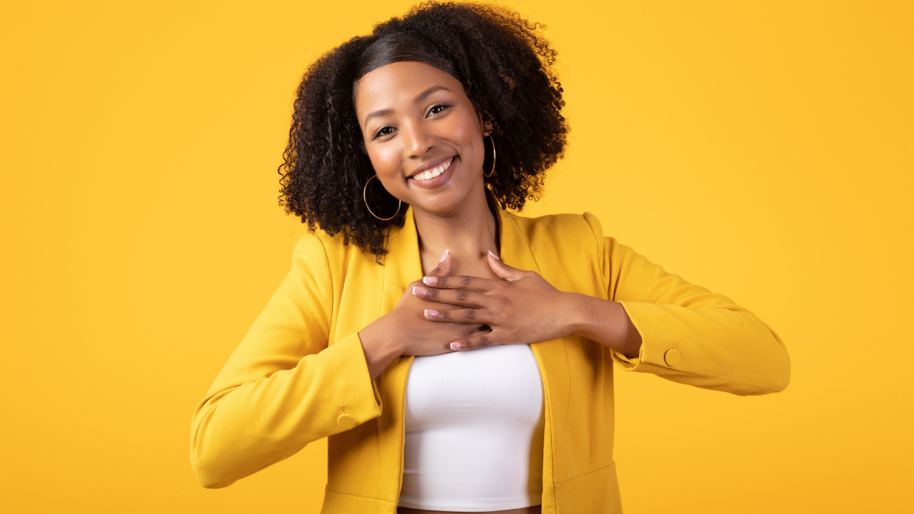 Mulher negra, com cabelo crespo, sorrindo com as mãos no coração usando blusa amarela em um fundo amarelo.