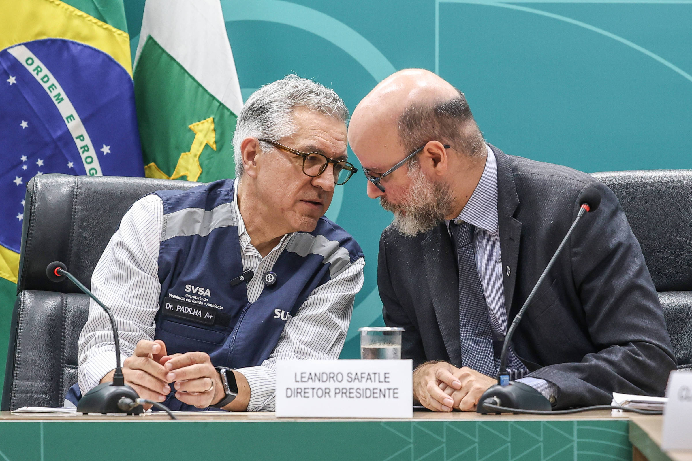 Leandro Safatle, diretor-presidente, conversa com assessor durante reunião em sala oficial. Bandeiras do Brasil e de estado ao fundo.