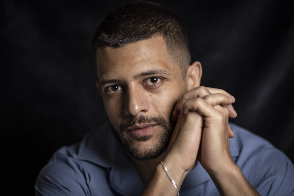 Homem de pele clara e cabelo curto olha diretamente para a câmera com as mãos entrelaçadas próximas ao rosto. Ele veste camisa azul e está contra fundo escuro.