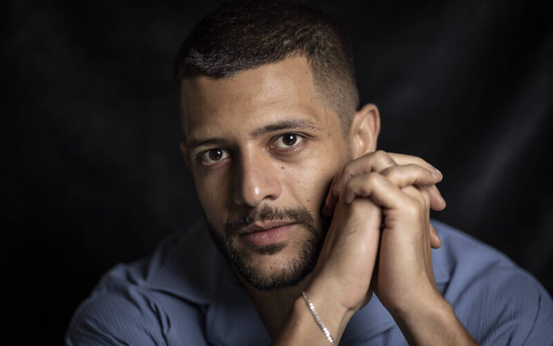 Homem de pele clara e cabelo curto olha diretamente para a câmera com as mãos entrelaçadas próximas ao rosto. Ele veste camisa azul e está contra fundo escuro.