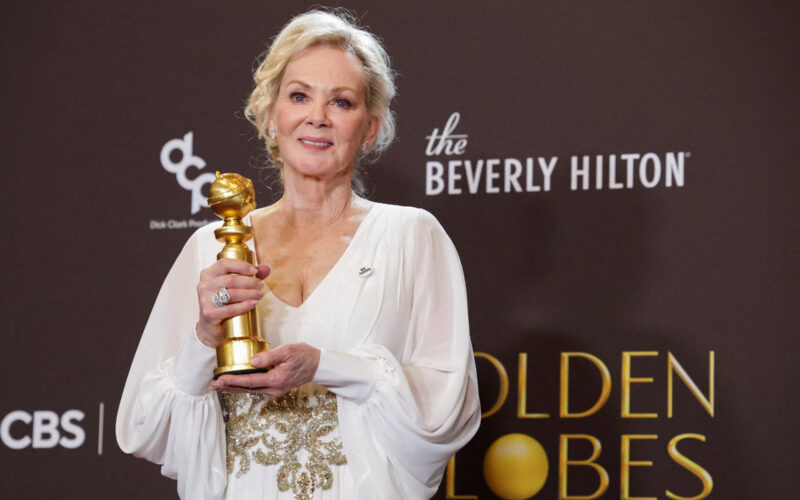 Mulher loira em vestido branco com detalhes dourados segura estatueta do Globo de Ouro em frente a fundo com logos do Beverly Hilton e Golden Globes.