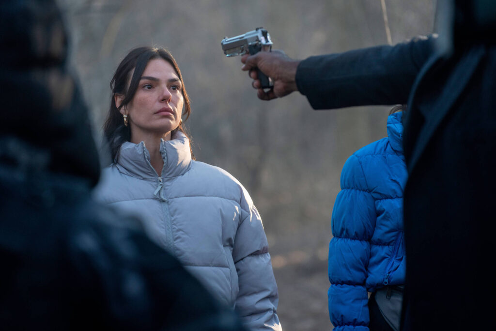 Mulher de casaco cinza claro está parada olhando para frente enquanto um homem aponta uma arma de fogo em sua direção. Ao fundo, outra pessoa veste casaco azul. Cena ocorre ao ar livre em ambiente com vegetação desfocada.