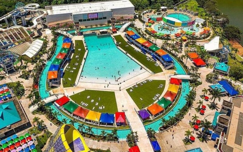 Vista aérea de parque aquático com piscina central grande em formato hexagonal, cercada por tendas coloridas e áreas gramadas com espreguiçadeiras. Ao redor, toboáguas, piscina circular menor, espreguiçadeiras com guarda-sóis coloridos e caminhos pavimentados. Vegetação e lago ao fundo.