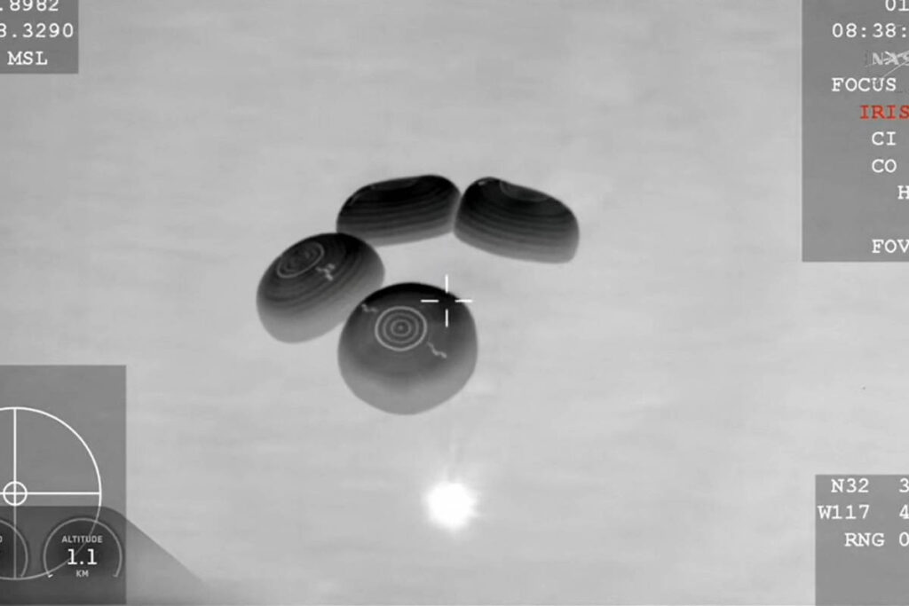 Tela em preto e branco mostra quatro objetos voadores com formato oval e listras, vistos por sistema de mira aérea com dados técnicos visíveis nas bordas da imagem.