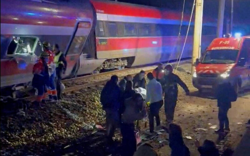 Trem com pintura vermelha, branca e azul parado nos trilhos à noite. Pessoas e equipes de resgate estão próximas ao trem e a uma ambulância com luzes ligadas.