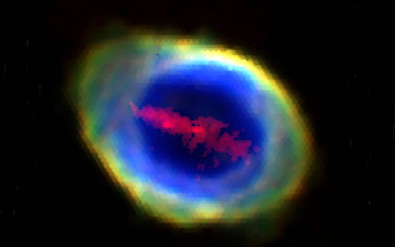 Nebulosa planetária com núcleo azul intenso cercado por anel difuso em tons de verde, amarelo e vermelho. Fundo preto com pontos de estrelas distantes.