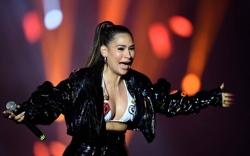 Cantora com cabelo preso em rabo de cavalo usa jaqueta preta brilhante e top branco estampado, segurando microfone com os braços abertos em palco iluminado por luzes alaranjadas e vermelhas desfocadas ao fundo.