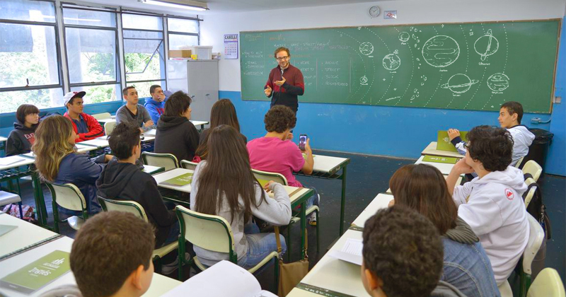 Sala de aula com professor falando em frente à lousa e alunos sentados acompanhando a apresentação