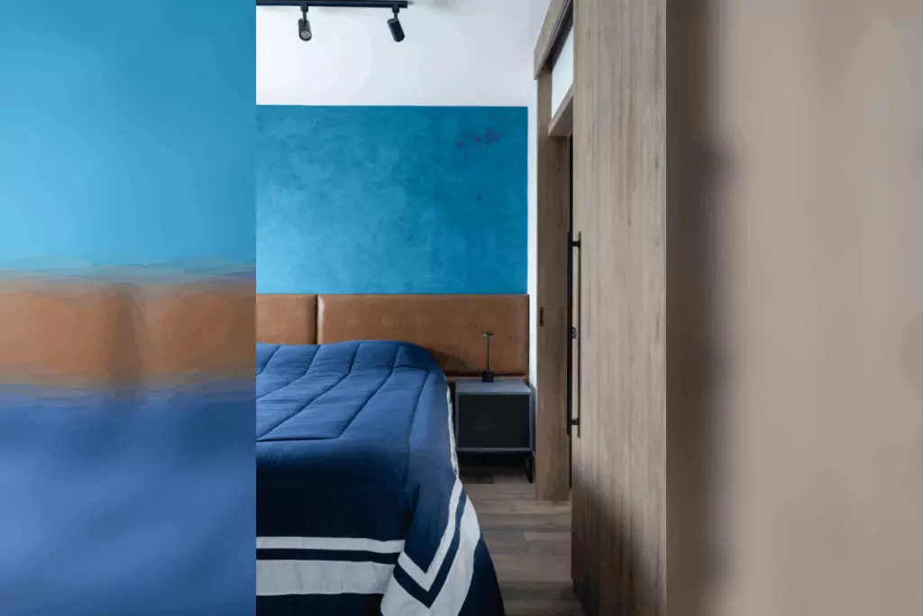 Quarto com cama posicionada junto à parede azul, criado-mudo lateral e porta de madeira