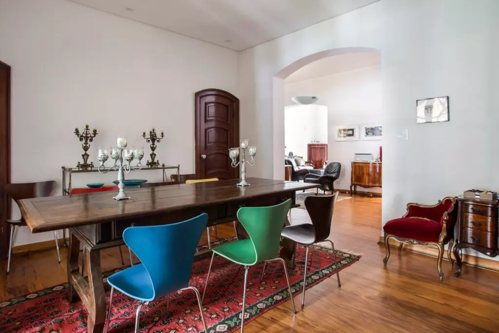 Sala de jantar com mesa de madeira, cadeiras coloridas, tapete e aparadores distribuídos pelo ambiente
